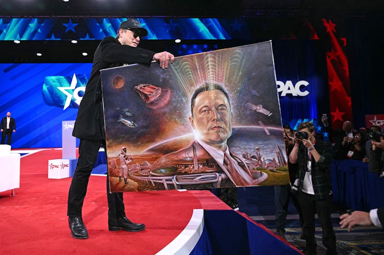 A white man with a dark hat and dark glasses wears black clothing and holds a large painting of himself on a stage.