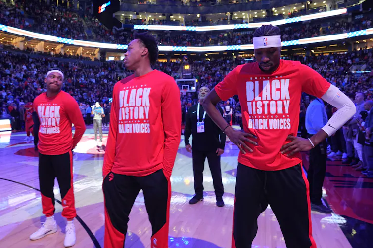 Dos hombres negros con mangas largas rojas se miran mientras están parados en una cancha de baloncesto frente a un estadio completo