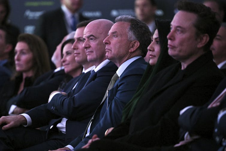 A row of middle-aged white men sit in a crowd of people