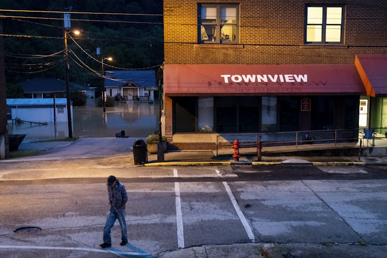 A man walks from a store with lighted rooms above it. In the background, homes are flooded.