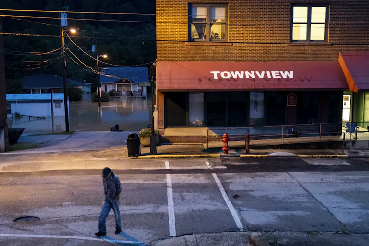 A man walks from a store with lighted rooms above it. In the background, homes are flooded.