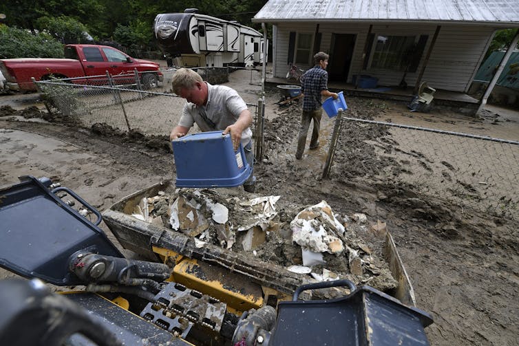 Two men dump buckets of ruined wallboard removed from a home. The yard they are walking through is filled with mud.
