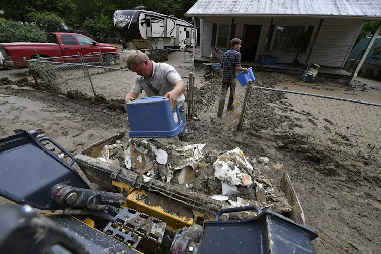 Two men dump buckets of ruined wallboard removed from a home. The yard they are walking through is filled with mud.