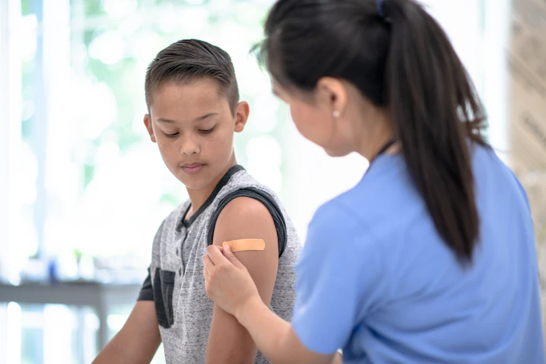 A nurse in blue scrubs places a band-aid on a young boy's arm after giving him a shot.