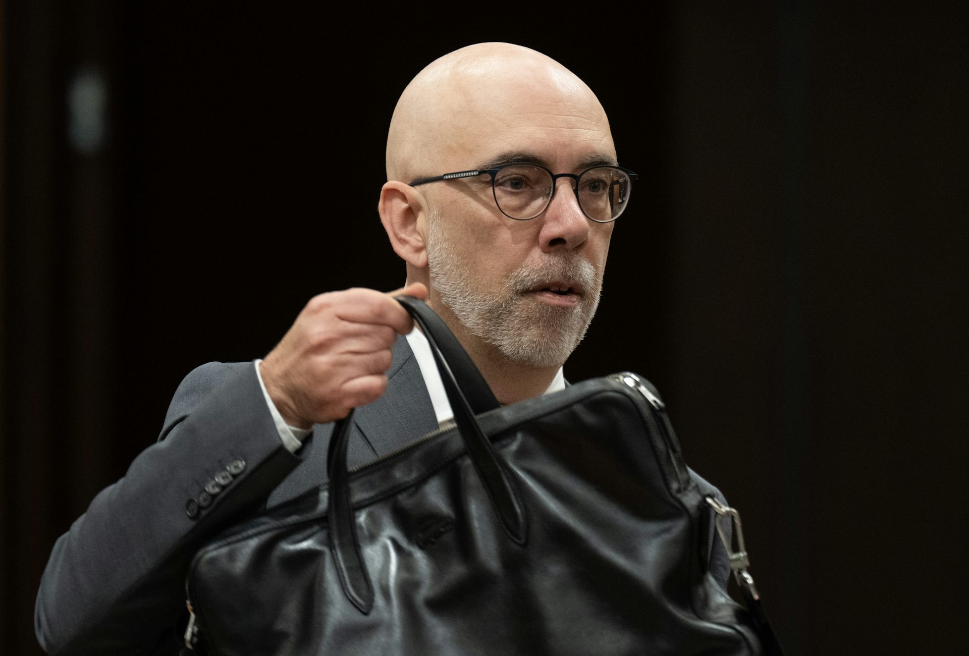 A bald white man wearing glasses and brown suits takes a black brief case