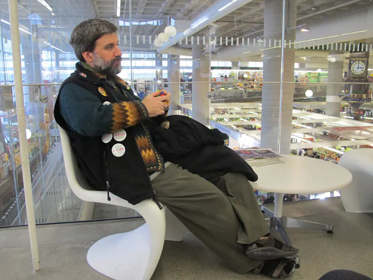 A man with greying hair and beard sits on a chair, books on shelves are seen in the background