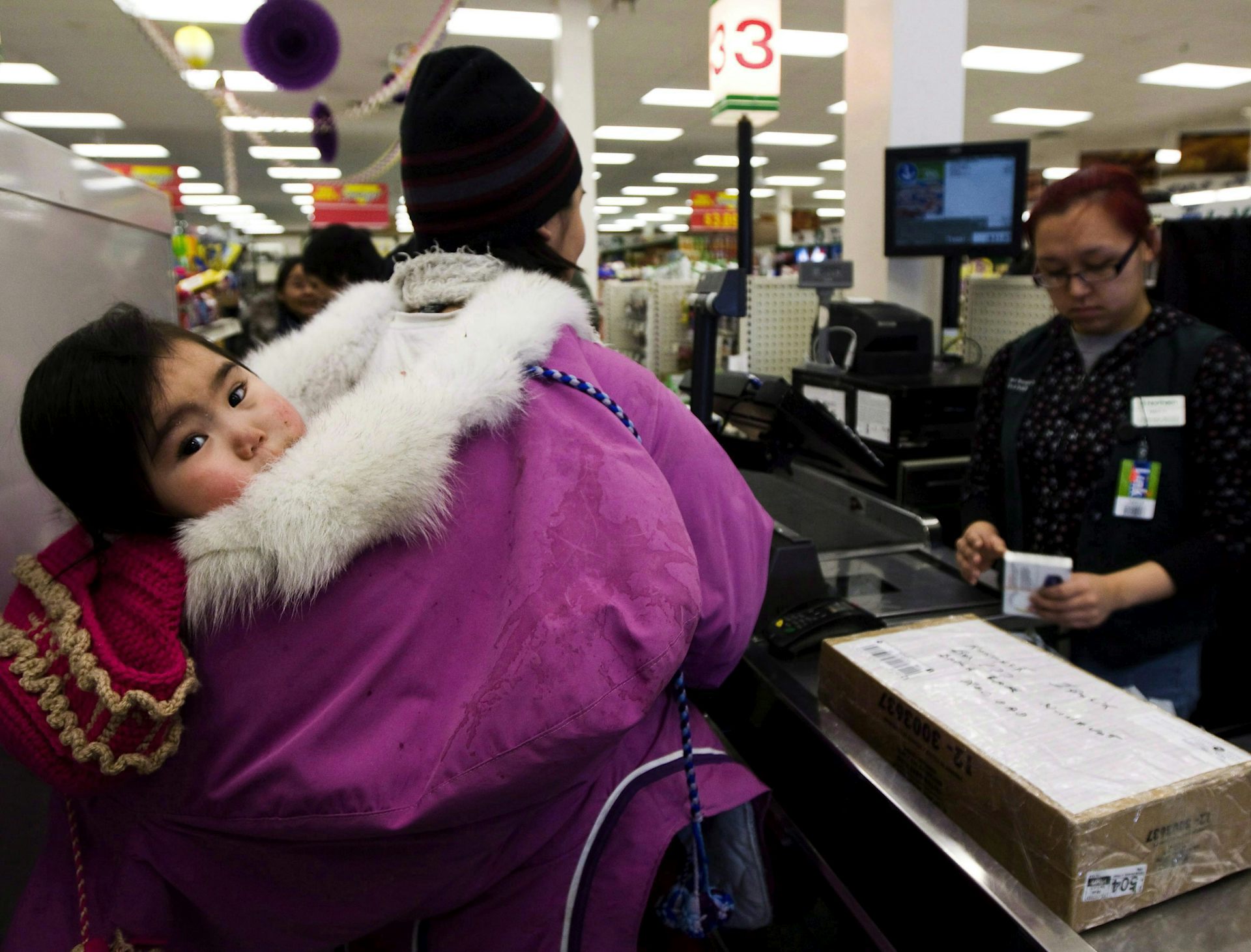 An Inuit woman wearing a purple parka carries a baby on her back. She stands at a grocery till with another woman behind the counter