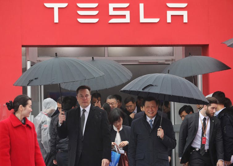 Men with umbrellas stand underneath a sign saying 'Tesla'