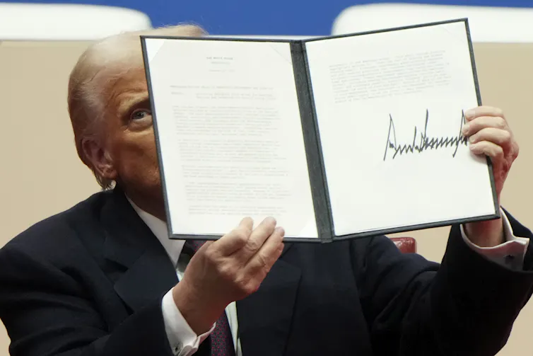 An old man in a dark suit with orange-tinted skin and yellowish-white hair holds up a document with an oversized signature at the bottom.