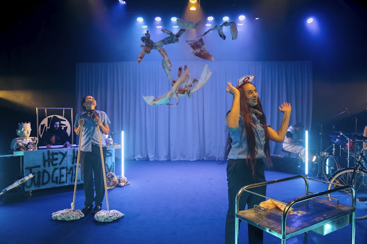 Two women stand on stage with a strange variety of props.