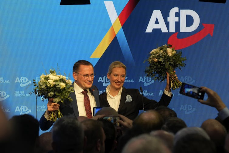 Man and woman in suits and with flowers standing on a stage waving to a crowd.
