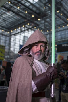 A man in a hooded brown cloak and dark leather gloves stands inside a tall atrium, holding a blue-green pole.