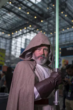 A man in a hooded brown cloak and dark leather gloves stands inside a tall atrium, holding a blue-green pole.