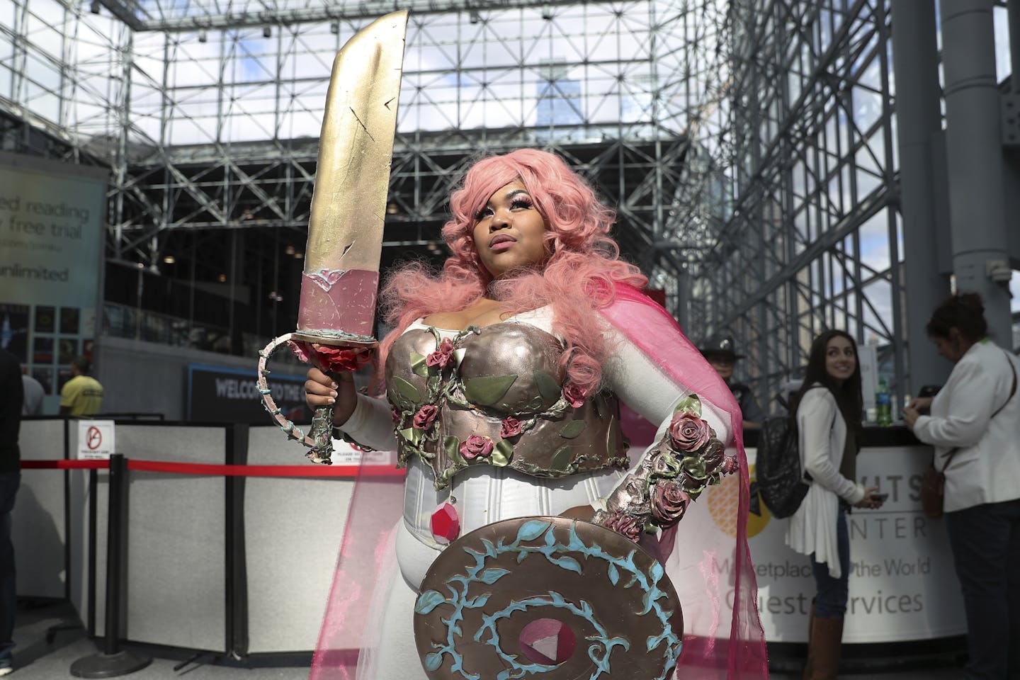 A woman holds a large sword as she poses in a pink wig, chest armor, a shield and a white-and-pink dress.