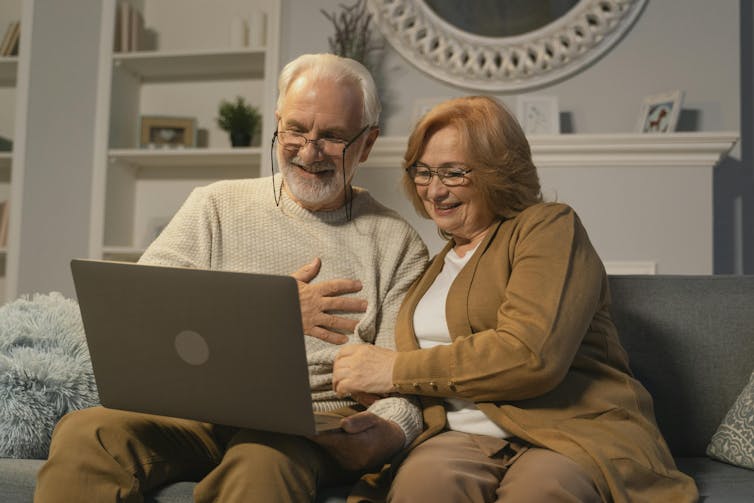 An elderly couple sit on a couch and smile while looking at a laptop screen.