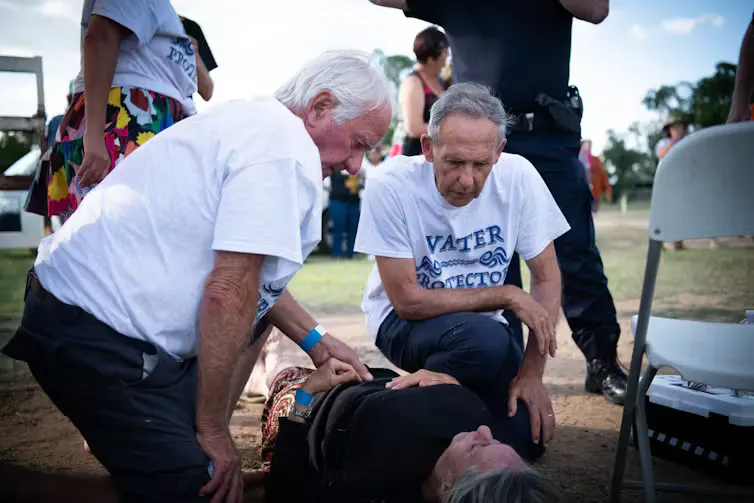 men kneeling around a woman lying injured on the ground.