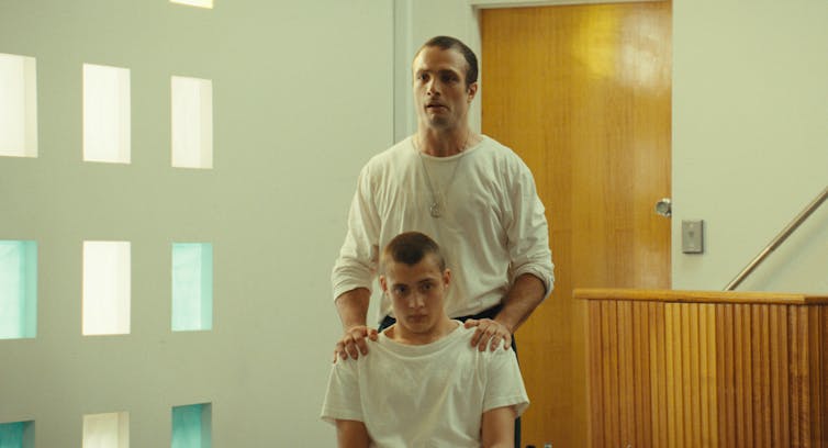 Film still: a man stands behind a man in a chair, hands on his shoulders.