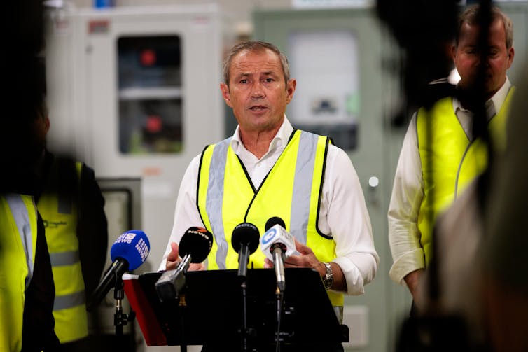 Male politician in high-vision vest speaks to the media at a podium.