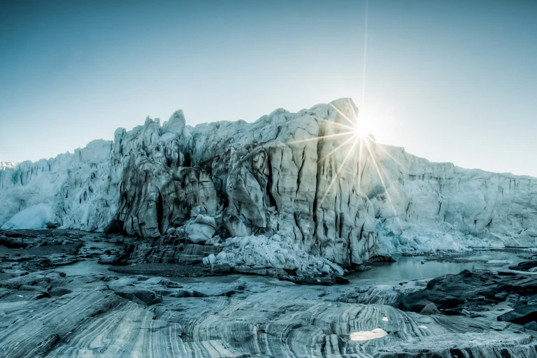 Photo of a glacier looming over rocks with the sun behind.