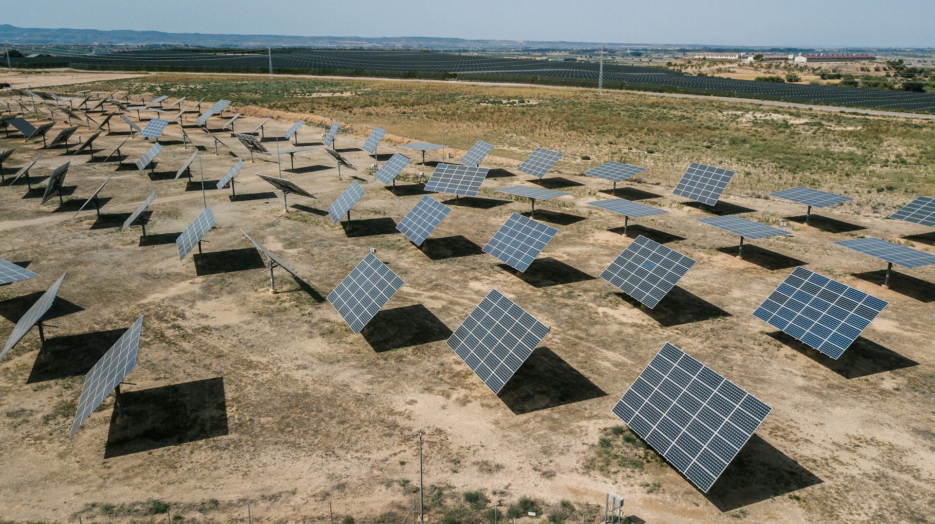 Rows of solar panels are located on a large field of light brown earth on a sunny day.