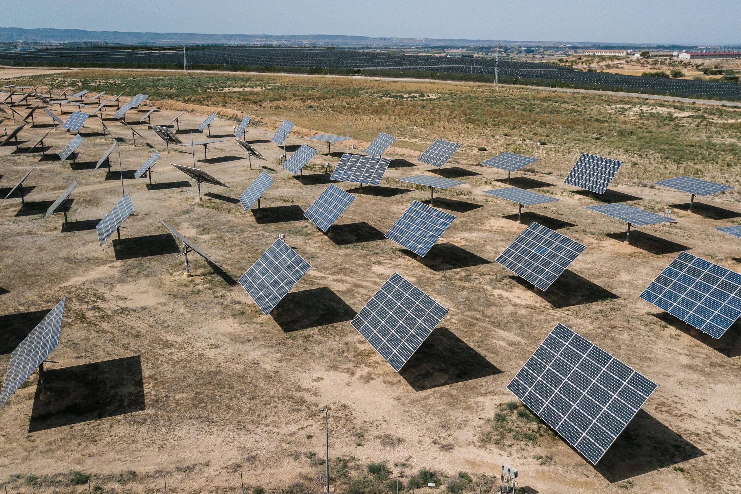 Rows of solar panels are located on a large field of light brown earth on a sunny day.