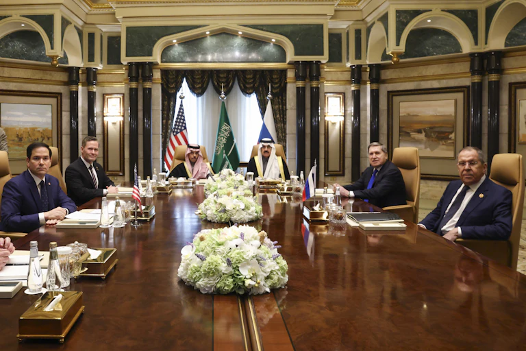 Seven men sit around a large wooden table in an ornate room. One man in the background records the meeting with a camera.