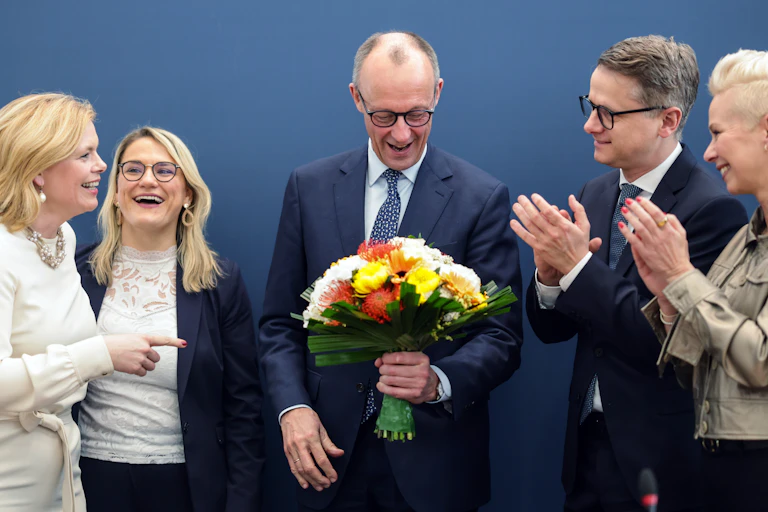 Friedrich Merz smiles with a bunch a flowers while people around him clap.