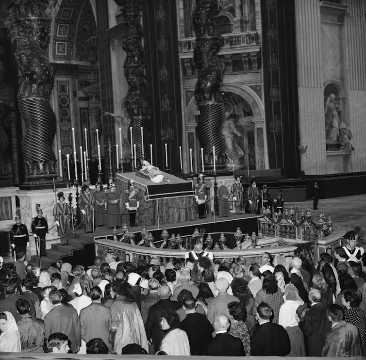 People walking around a coffin lying in the dome of a large hall with intricate carvings.