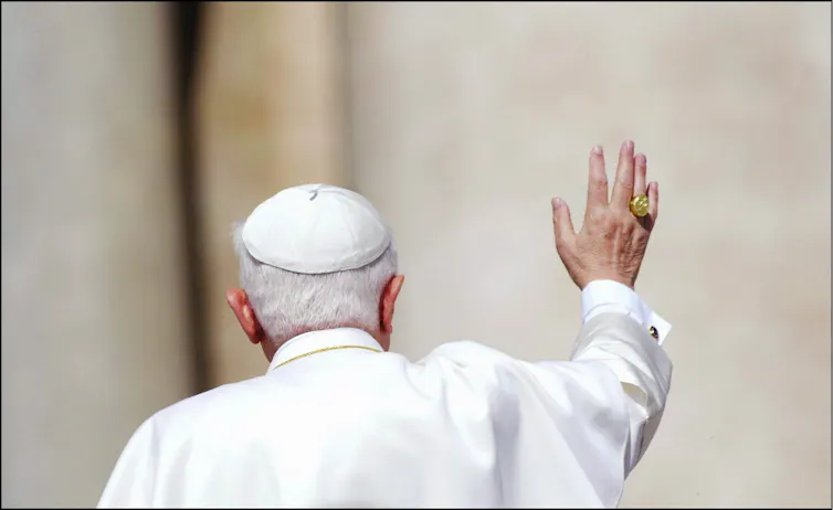 The back of a man wearing a white robe and cap, waving with his hand, which bears a golden ring.