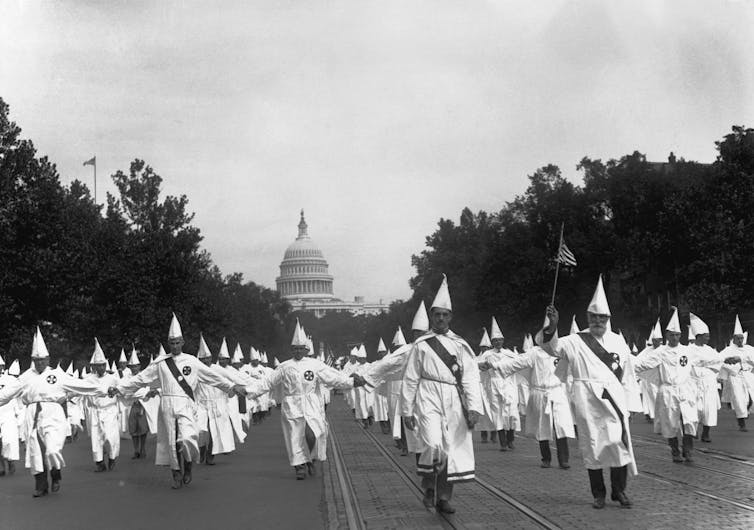 A large group of men wearing white gowns and white pointed hats walk in uniform, with a large dome building behind them in a black-and-white photo.