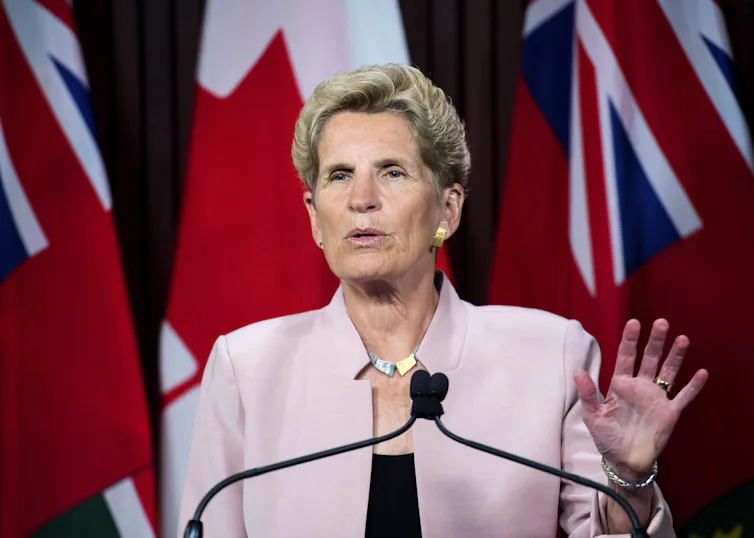 A middle-aged white woman with short blonde hair speaks into a microphone on a podium while standing in front of the Canadian and Ontario flags