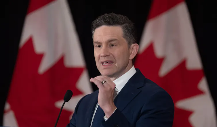 Hombre blanco de mediana edad con cabello corto y oscuro habla en un micrófono mientras está de pie en la parte delantera de las banderas canadienses
