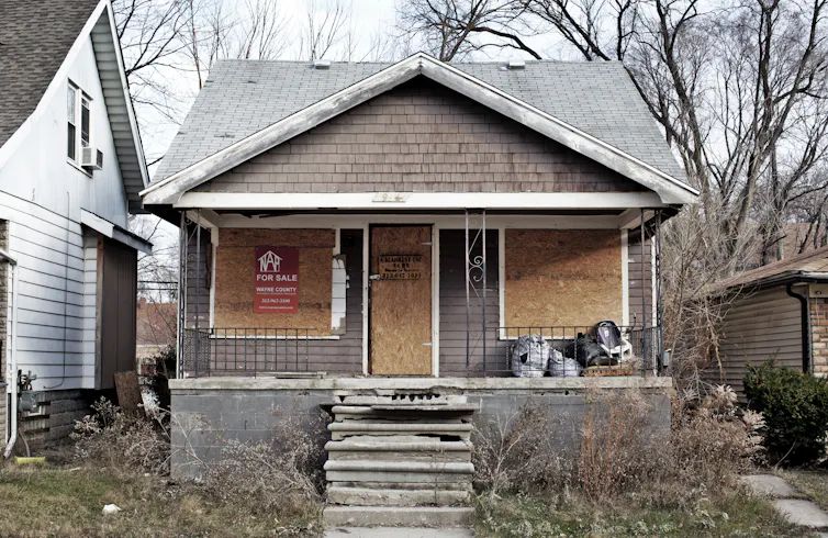 A grey house with chipped steps, shuttered windows and overgrown weeds in the yard. There is a for sale sign and it is clearly in a state of disrepair.