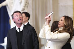 A man in a dark suit looks up with his mouth open while a woman dressed in white takes a photo of a room with her phone