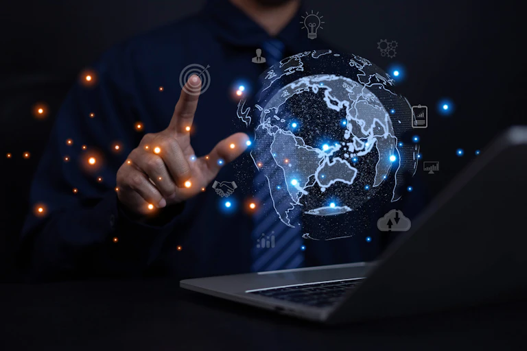 A holographic image of the earth floats above an open laptop. A person, seen from the neck-down, touches an image of a circle floating above the earth