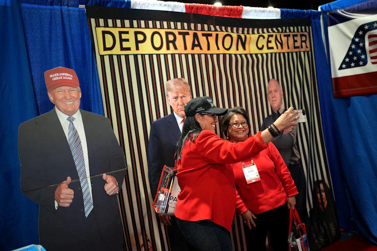 Duas mulheres de vermelho seguram um telefone celular e sorriem. Elas estão em frente a um cenário com listras amarelas e pretas que diz “Centro de deportação”. Há recortes de Donald Trump em frente a ele.