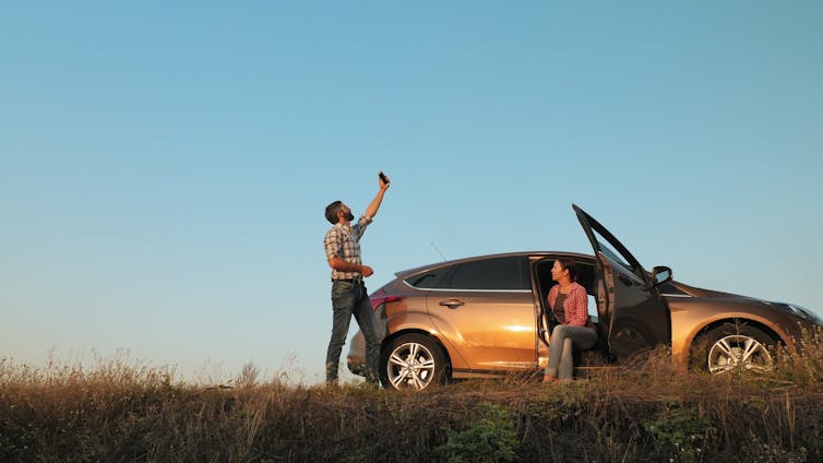 A man holds his mobile phone to the sky, while a woman sits in a broken-down car on the side of a remote road.