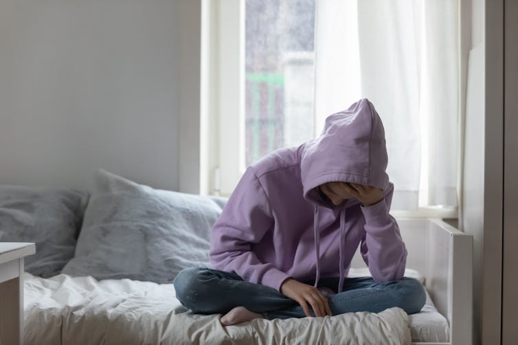 An unhappy-looking child in a purple hoodie sits on her bed with her head in her hands.