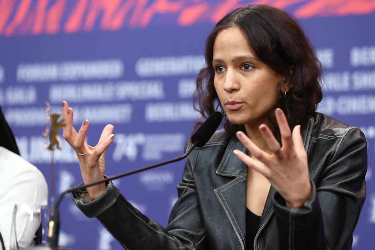 A woman in a denim jacket gestures with both hands, fingers splayed, as she talks into a microphone at a desk.