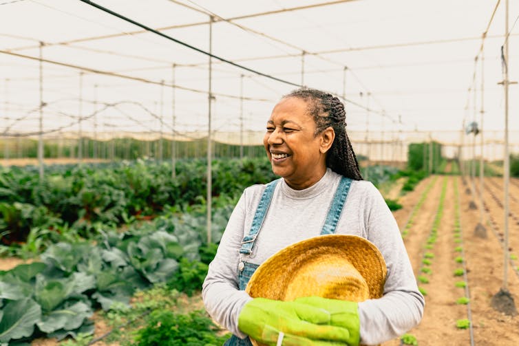 A woman farmer at a cabbage farm clutches a straw hat with both hands and smiles.