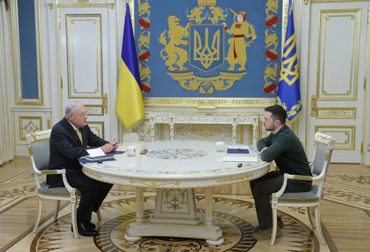Volodymyr Zelensky and Keith Kellogg sitting opposite each other at a large table.