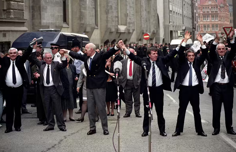The six Birmingham Six men stand in the street behind microphones with their arms raised.
