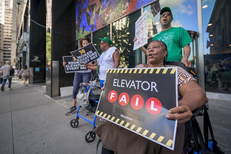 A group of people stand and sit, in a wheelchair, on a street. They hold black and white signs. One of them says 'Elevator Fail.'