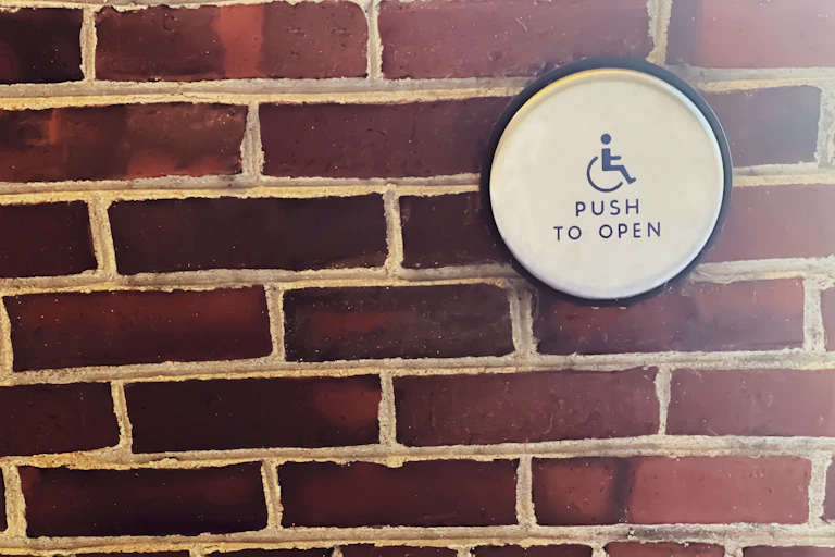 A brick wall has a white circular sign on it with a picture of a wheelchair that says 'Push to open.'