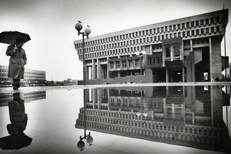 Man holding an umbrella walks across a plaza in front of a large, imposing concrete building.
