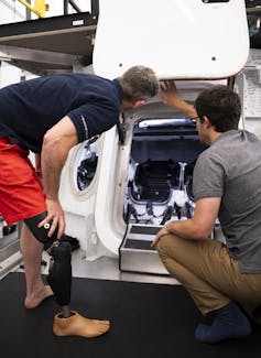 A man with a prosthetic leg leans over next to mockup of a space capsule with the hatch open, another man kneels beside him.