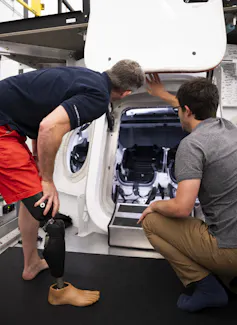 A man with a prosthetic leg leans over next to mockup of a space capsule with the hatch open, another man kneels beside him.