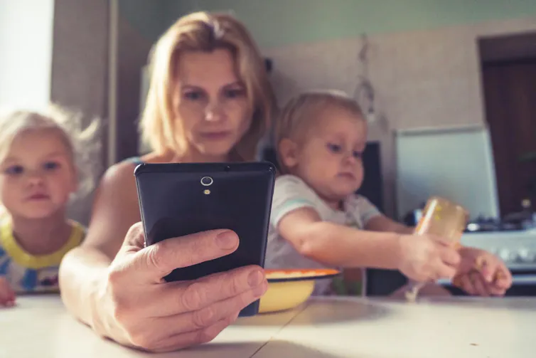 A woman looks at a cellphone while holding a child in her lap at a kitchen table. Another child looks on