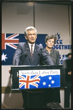 Bob Hawke stands addressing the media with an Australian flag in the background.