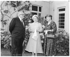 Black and white photo of a man in a suit standing with two women in dresses in a garden.
